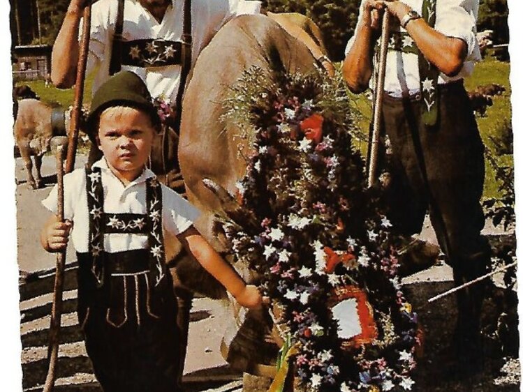 Two men in traditional Alpine costumes carrying a floral wreath, accompanied by a young child, in a scenic outdoor setting. | © Postkarte/Milz/1952