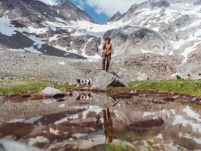 Snow-capped mountains, a person standing on a rock, a small dog, and a calm reflection pool in a grassy landscape. | © Urlaub am Bauernhof / Daniel Gollner