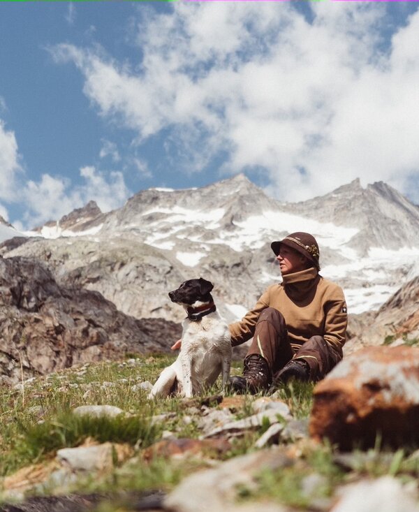 A person sitting on a grassy slope with a dog, surrounded by snow-capped mountains and cloudy sky in a scenic outdoor setting. | © Urlaub am Bauernhof / Daniel Gollner