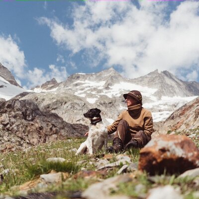 A person sitting on a grassy slope with a dog, surrounded by snow-capped mountains and cloudy sky in a scenic outdoor setting. | © Urlaub am Bauernhof / Daniel Gollner