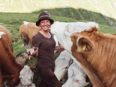 Smiling woman in hat standing among cows in a grassy field, holding a milking device. | © Urlaub am Bauernhof / Daniel Gollner