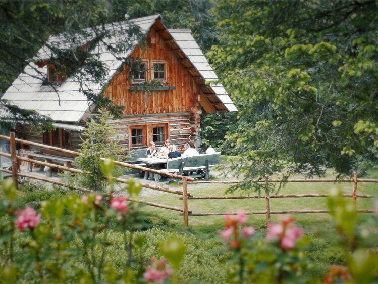Malerisches Holzhaus umgeben von Bäumen und Blumen, auf einer Terrasse sitzen Menschen und genießen die Natur.