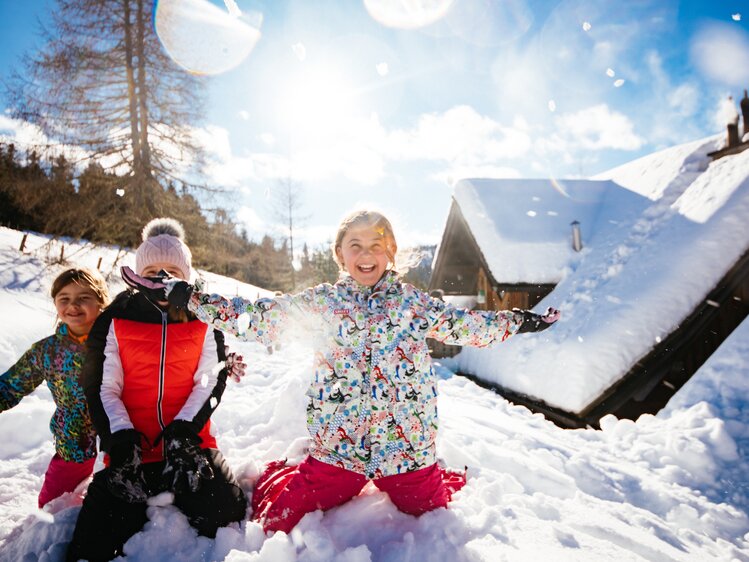 Fröhliche Kinder in bunten Winterjacken und Hosen spielen ausgelassen im tiefen Schnee vor einer verschneiten Berglandschaft, die Arme ausgebreitet vor Freude. | © Urlaub am Bauernhof Kärnten / Daniel Gollner