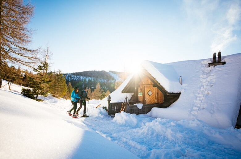 Ein schneebedecktes Berghaus in einer winterlichen Landschaft. Zwei Menschen in Sportkleidung genießen die Winterlandschaft. Die Sonne scheint durch die Bäume und wirft einen warmen Lichtschein auf die Szenerie. | © Daniel Gollner