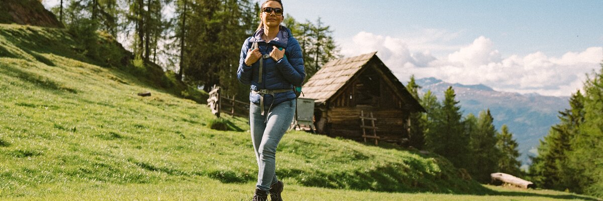 Eine Wanderin in Jeans und Jacke steht auf einer grünen Wiese vor einer Holzhütte, umgeben von Bäumen und Berglandschaft in der Ferne. | © Urlaub am Bauernhof Kärnten / Daniel Gollner