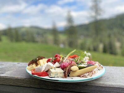 Eine Auswahl an Wurst, Käse, Tomaten, Gewürzen und anderen Lebensmitteln auf einem holzigen Untergrund, umgeben von einer grünen, bergigen Landschaft. | © Urlaub am Bauernhof Kärnten / Jasmin Kreulitsch