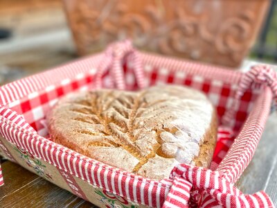 Hausgebackener Brotlaib in dekorativer Holzschale, umwickelt mit karierter rot-weißer Serviette, eingebettet in eine blumige Umgebung. | © Urlaub am Bauernhof Kärnten / Jasmin Kreulitsch