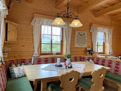 Gemütliches Holzhaus mit rustikalem Interieur, großer Esstisch mit bestickten Sitzbänken, traditionelle Details wie Vorhänge und Kerzen, Blumen und ein Schild mit Spruch, eine gemütliche und einladende Atmosphäre. | © Urlaub am Bauernhof Kärnten / Jasmin Kreulitsch