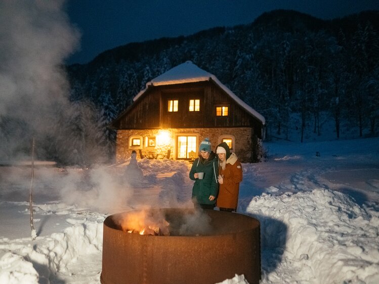 Eine verschneite Hütte bei Nacht, mit warm erleuchteten Fenstern und einem lodernden Feuer im Freien, an dem zwei Menschen stehen und sich wärmen. | © Urlaub am Bauernhof / Daniel Gollner