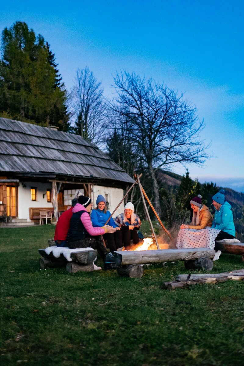 Eine Gruppe von Personen sitzt bei Nacht an einem Lagerfeuer vor einer traditionellen Holzhütte umgeben von Wald und Bergen. Die Szene erscheint gemütlich und einladend. | © Urlaub am Bauernhof Kärnten/ Daniel Gollner