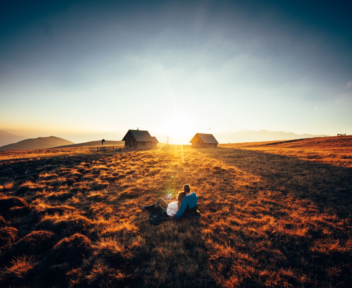 Ein romantischer Sonnenuntergang auf der Alm in Kärnten | © Urlaub am Bauernhof Kärnten/ Daniel Gollner Paar sitzt vor der Hütte im Gras und genießt den Sonnenuntergang auf der Alm | © Urlaub am Bauernhof Kärnten/ Daniel Gollner