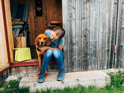 Mädchen und Hund sitzen in der Almhüttentüre | © Urlaub am Bauernhof Kärnten / Daniel Gollner A woman sitting on a wooden porch, hugging a brown dog in front of a weathered wooden wall. | © Urlaub am Bauernhof Kärnten / Daniel Gollner