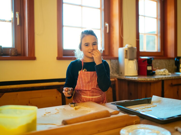 Eine junge Frau in einer kariertemSchürze bereitet etwas in einer Küche vor, umgeben von Küchenutensilien und einem hell erleuchteten Raum mit alten Holzfenstern. | © Urlaub am Bauernhof Kärnten / Daniel Gollner