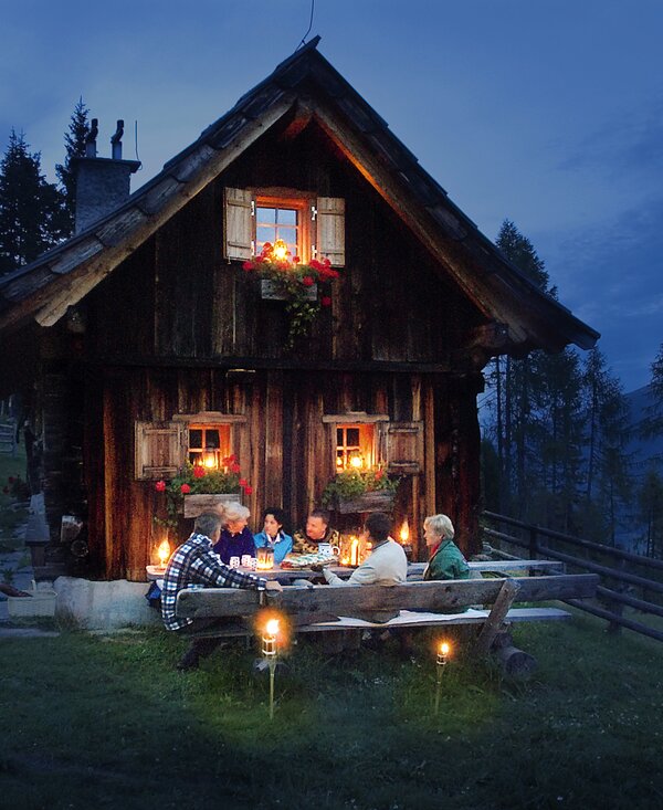 Cozy wooden cottage with lighted windows, festive decorations, and a group of people enjoying a meal outdoors on a starry night. | © Urlaub am Bauernhof Kärnten/ Ernst P. Prokop