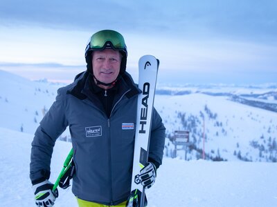 Franz Klammer | © BRM / Mathias Prägant Person wearing ski attire, holding skis, standing on snowy mountain against cloudy sky. | © BRM / Mathias Prägant