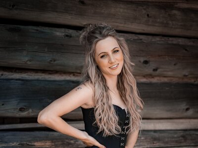 Melissa Naschenweng | © Lambauer Entertainment / Christoph Hatheuer Woman with long gray hair wearing a black fringe top and pink metallic shorts, posing against a rustic wooden wall. | © Lambauer Entertainment / Christoph Hatheuer