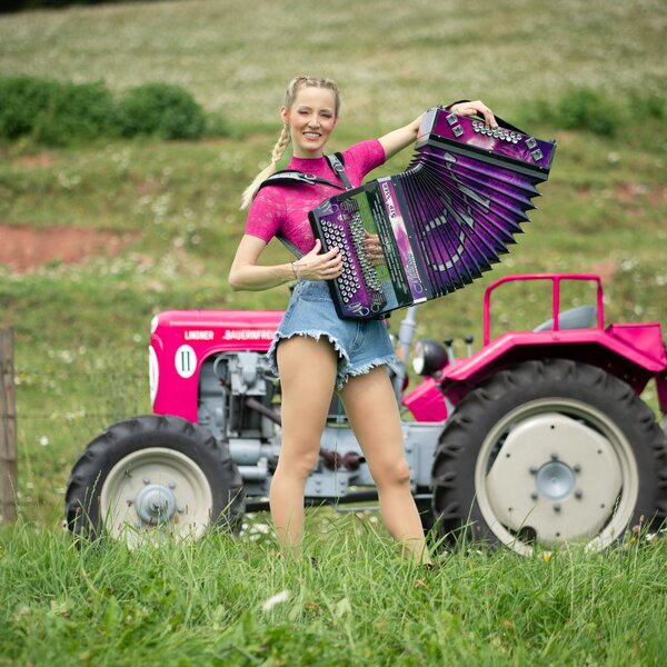Smiling woman posing with colorful accordion on a red tractor in a grassy field. | © Lambauer Entertainment / Christoph Hatheuer
