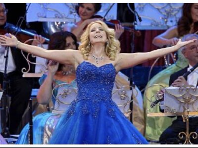 Eine Frau im prachtvollen blauen Ballkleid mit ausgebreiteten Armen und strahlendem Lächeln, umgeben von einer Orchesterkulisse. | © Urlaub am Bauernhof / Schell