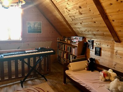 Gemütliches Dachzimmer mit Klavier, Bücherregal und Bett, umgeben von Holzpaneelen und Lichteinfall durch Fenster. Ruhige, heimelige Atmosphäre. | © Urlaub am Bauernhof / Schell