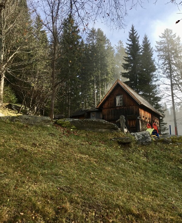 Ein baufälliges Holzhaus steht inmitten eines bewaldeten Berghanggeländes mit Moos und abgestorbenen Ästen. Im Vordergrund sind zwei Personen in Regenjacken zu erkennen. | © Urlaub am Bauernhof / Schell