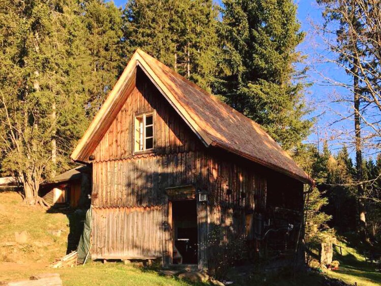 Ein altes, hölzernes Berghaus in einer idyllischen Waldumgebung mit hohen Bäumen, unter einem blauen Himmel. Das Gebäude wirkt rustikal und passt harmonisch in die natürliche Umgebung. | © Urlaub am Bauernhof / Schell