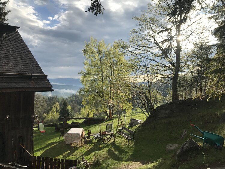 Eine gemütliche Außenanlage mit Blick auf bewaldete Hügel und einen bewölkten Himmel. Ein Holzhaus, Möbel und Gartengeräte ergeben eine ruhige, friedliche Szenerie umgeben von üppiger Vegetation. | © Urlaub am Bauernhof / Schell