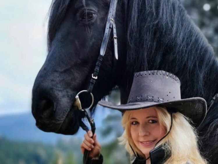 Eine Frau in Cowboyhut steht neben einem schwarzen Pferd in einer ländlichen Umgebung. | © Urlaub am Bauernhof / Schell