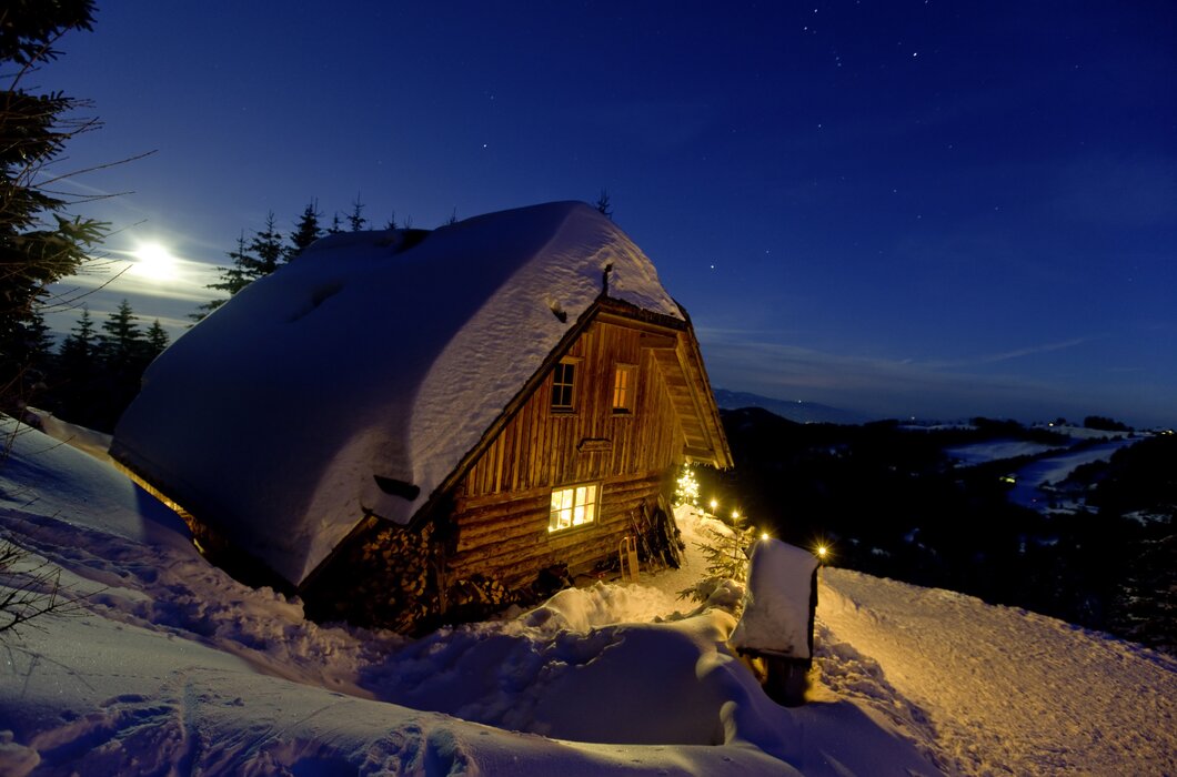Eine verschneite Berghütte mit beleuchteten Fenstern und einer schneebedeckten Umgebung unter einem sternenübersäten Nachthimmel. | © Urlaub am Bauernhof/ Tom Lamm
