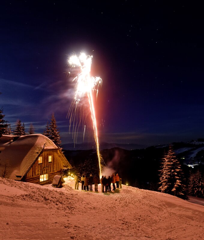 Eine Menschengruppe versammelt sich vor einer beleuchteten Hütte, während oben am Nachthimmel bunte Feuerwerksexplosionen zu sehen sind. Die Szene vermittelt eine winterliche Feststimmung in einer bergigen Landschaft. | © Urlaub am Bauernhof/ Tom Lamm