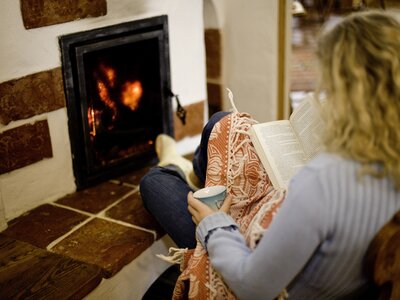 Frau wärmt sich am Ofen auf der Alm | © Urlaub am Bauernhof/ Tom Lamm Eine Person sitzt gemütlich auf dem Boden vor einem Kamin, ein Buch in der Hand. Das warme Feuer bringt ein behagliches Ambiente hervor. | © Urlaub am Bauernhof/ Tom Lamm