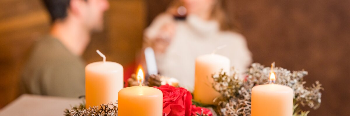 Adventskranz mit brennenden Kerzen, Tannengrün, Zapfen und Rosen auf einem Holztisch. Ein Paar in unklarer Umgebung blickt darauf.