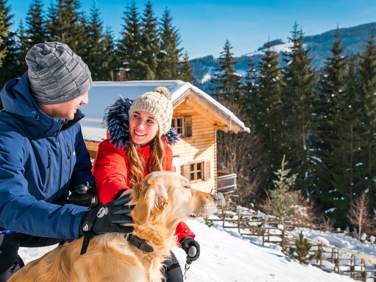 Couple enjoying a snowy winter day with their golden retriever in front of a rustic wooden cabin in the mountains. | © Michael Stabentheiner