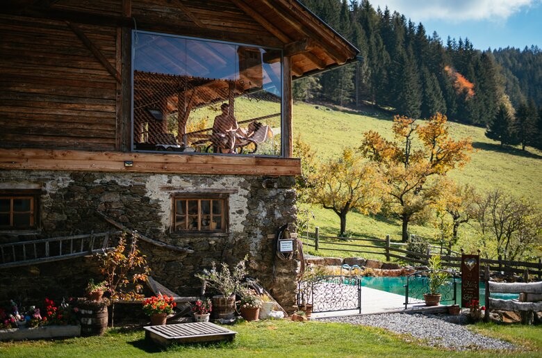Urige Hütte mit Holzfassade und steinernem Erdgeschoss inmitten einer Herbstlandschaft mit leuchtenden Bäumen und Pool. Rustikales Ambiente lädt zum Entspannen ein. | © Urlaub am Bauernhof / Daniel Gollner
