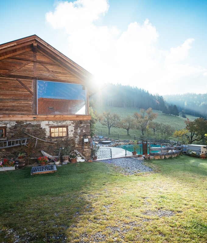 A rustic wooden chalet surrounded by lush greenery, with a grassy field, stone walls, and a view of forested hills in the distance. | © Urlaub am Bauernhof / Daniel Gollner