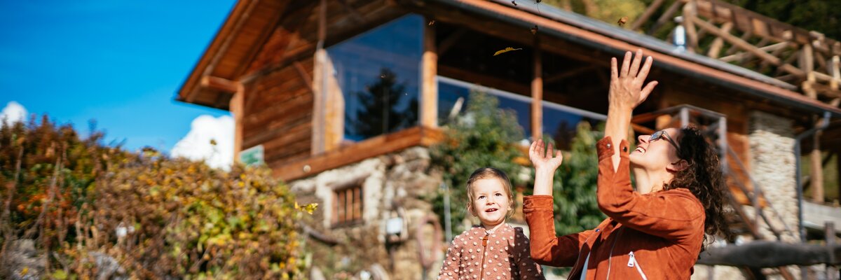 Eine Mutter und ihr Kind spielen im Freien, umgeben von herbstlichen Blättern, vor einer hölzernen Berghütte. Die Szene strahlt Naturnähe und Freude aus. | © Urlaub am Bauernhof / Daniel Gollner
