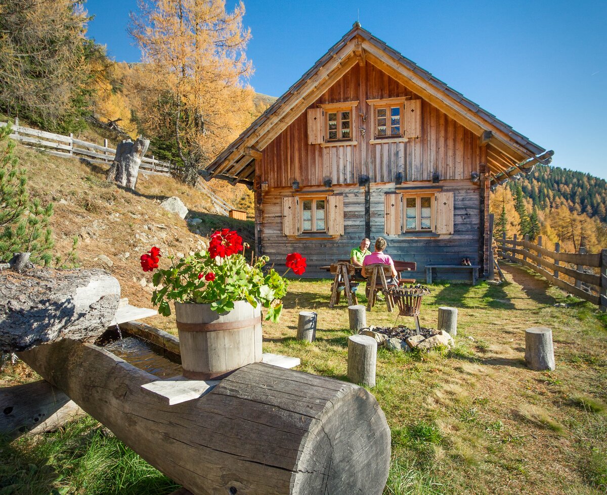 Traditionelles Holzhaus in malerischer Landschaft, umgeben von herbstlichen Bäumen und geschmückt mit Blumen. Gemütliche Sitzgelegenheiten laden zum Verweilen ein.