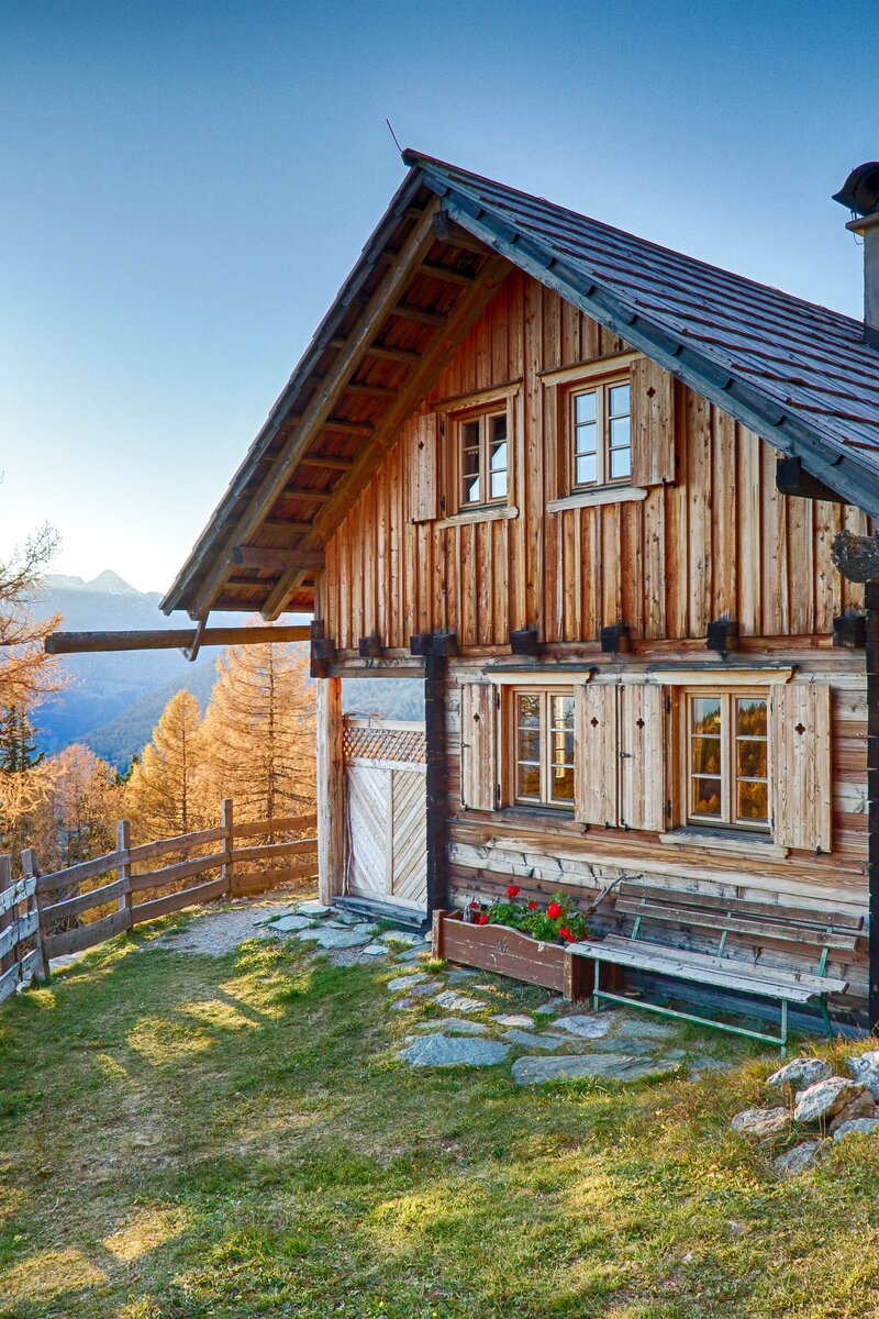Einsames, romantisches Berghaus mit Holzfassade, umgeben von buntem Herbstlaub und weiter Landschaft. Kleine Fenster, Kamin und geschwungenes Dach verleihen dem Gebäude rustikalen Charme.
