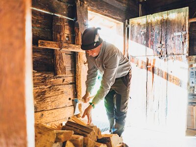 Beim Holzhacken auf der Alm | © Urlaub am Bauernhof Salzburg / Daniel Gollner Eine Person in einem karierten Hemd und Kappe bearbeitet Holzbretter in einer rustikalen Werkstatt mit Holzwänden und -konstruktionen. Im Hintergrund sind verschiedene Werkzeuge und Dekorationen zu sehen. | © Urlaub am Bauernhof Salzburg / Daniel Gollner