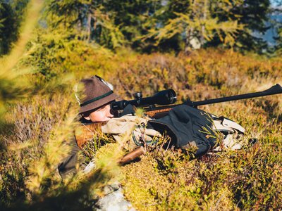 Jäger auf der Pirsch | © Urlaub am Bauernhof Salzburg / Daniel Gollner Eine Person in Tarnkleidung und Hut liegt mit einem Gewehr bewaffnet auf dem Boden, umgeben von herbstlicher Vegetation. | © Urlaub am Bauernhof Salzburg / Daniel Gollner