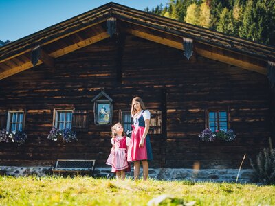 Mädchen im Dirndl vor der Almhütte | © Urlaub am Bauernhof Salzburg / Daniel Gollner Eine Frau und ein Mädchen in traditioneller bayerischer Tracht vor einer rustikalen Holzhütte mit Blumenbeeten und umgeben von berghafter Landschaft. | © Urlaub am Bauernhof Salzburg / Daniel Gollner