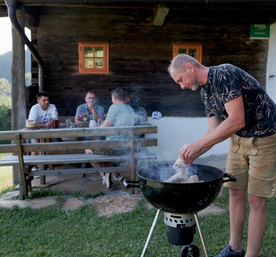Ein Mann heizt vor der Almhütte einen Griller an. Die restlichen Freunde sitzen im Hintergrund auf der Terrasse, reden miteinander und trinken gemeinsam. | © Urlaub am Bauernhof / Daniel Gollner