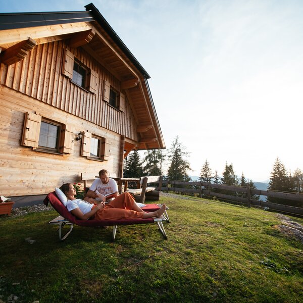 Wooden chalet with guests relaxing on the lawn overlooking a scenic view. | © Urlaub am Bauernhof Steiermark / Daniel Gollner