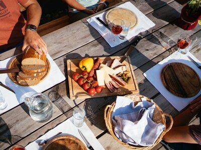 Gemütliches Holztisch-Picknick im Freien mit Brot, Käse, Wurst, Oliven und Gläsern für Wein oder Schnaps. Frische Früchte und Gemüse sowie Besteck und Servietten vervollständigen die Tischausstattung. | © Urlaub am Bauernhof Steiermark / Daniel Gollner