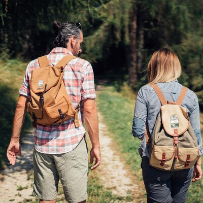 Zwei wandernde Personen mit Rucksäcken gehen auf einem Waldweg durch eine grüne Umgebung. Die Person vorne trägt ein kariertes Hemd, die andere Person eine Jacke mit Karo-Muster. | © Urlaub am Bauernhof Steiermark / Daniel Gollner 