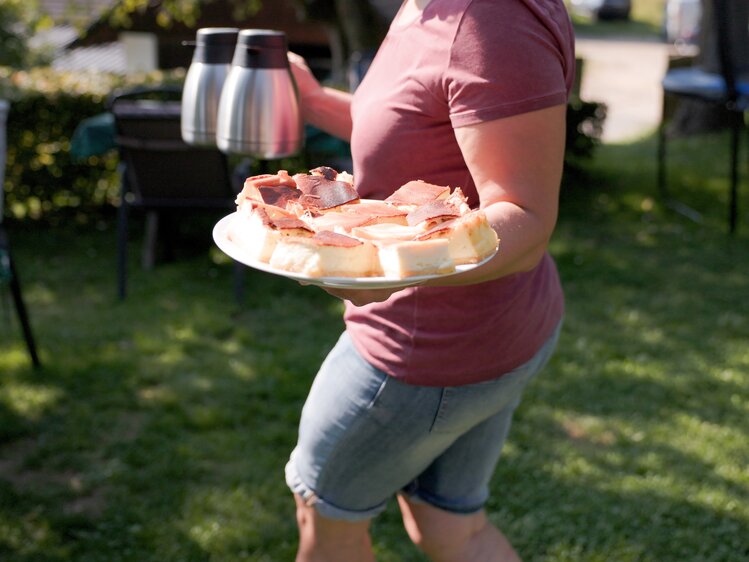 Person hält eine Platte mit gegrilltem Fleisch und Käse im Garten bei gutem Wetter.