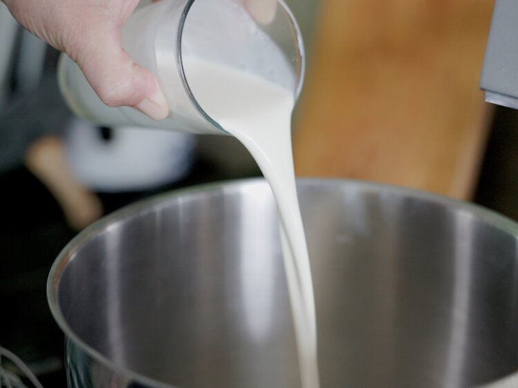 Jemand gießt eine Milch in eine Küchenmaschine, wahrscheinlich beim Backen oder Kochen.