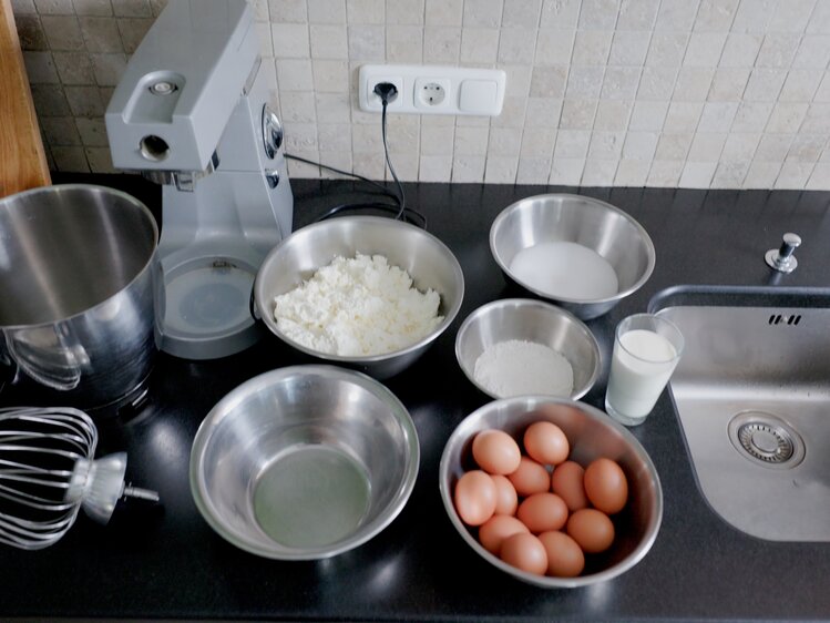 Zutaten und Küchengeräte auf einer schwarzen Arbeitsplatte, einschließlich Eier, Milch, Mehl, Zucker, Wasser, Rührgerät und Mixer