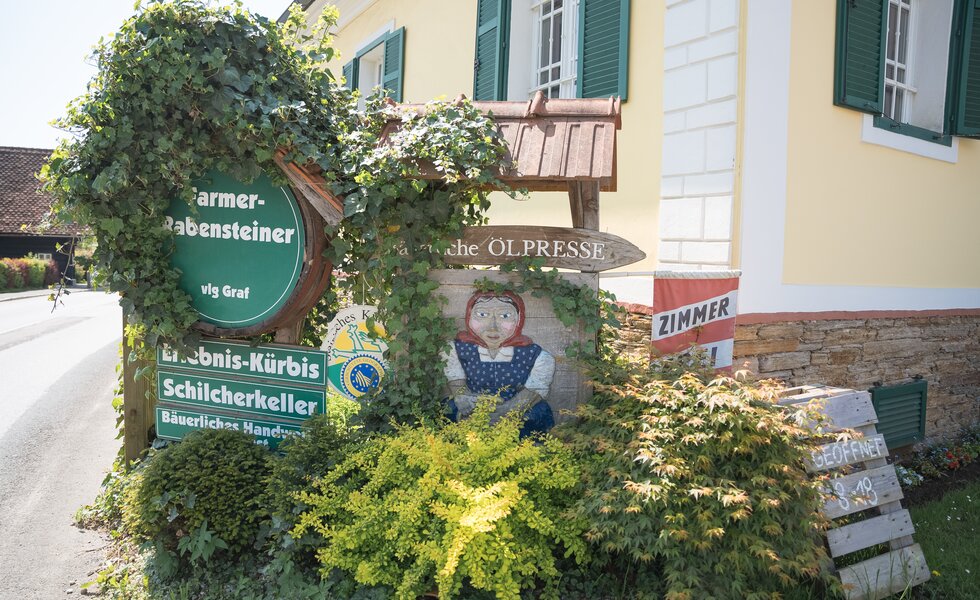 Typische ländliche Häuserfassade mit Rankgewächsen, Holzfässern und Schildern lokaler Anbieter landwirtschaftlicher Produkte. Bunte, idyllische Dorfszene mit einem Märchenfigur-Motiv an der Wand. | © Urlaub am Bauernhof / Daniel Gollner