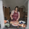 Eine Frau in einer Kochschürze sitzt an einem Küchentisch und bereitet ein Gericht zu, umgeben von Lebensmitteln wie Karotten, Zwiebeln und Schinkenwürfeln.