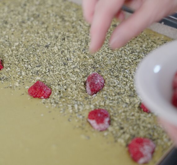 Eine Hand füllt frische Himbeeren in eine Schüssel mit zerkleinerter grüner Masse, die möglicherweise zu Kräutern oder Müsli verarbeitet wird.
